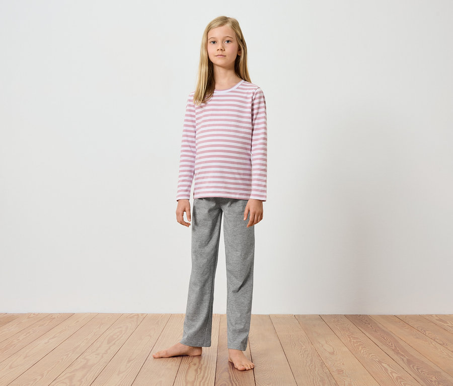 Młoda dziewczyna stoi boso na drewnianej podłodze na tle białej ściany. Ma na sobie bluzkę z długim rękawem w różowo-białe paski i szare spodnie.