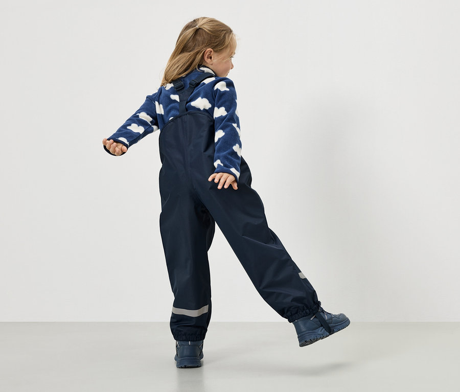 Dziewczynka w granatowych spodniach przeciwdeszczowych na szelkach i niebieskim swetrze w chmury, widziana od tyłu.