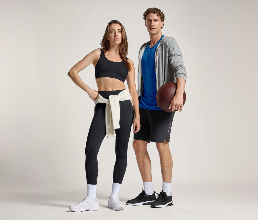 Kobieta w czarnym biustonoszu sportowym i legginsach sportowych „ActivePower” ze swetrem zawiązanym w pasie stoi obok mężczyzny w niebieskiej koszulce, szarej bluzie z kapturem i szortach, trzymającego piłkę do futbolu amerykańskiego.