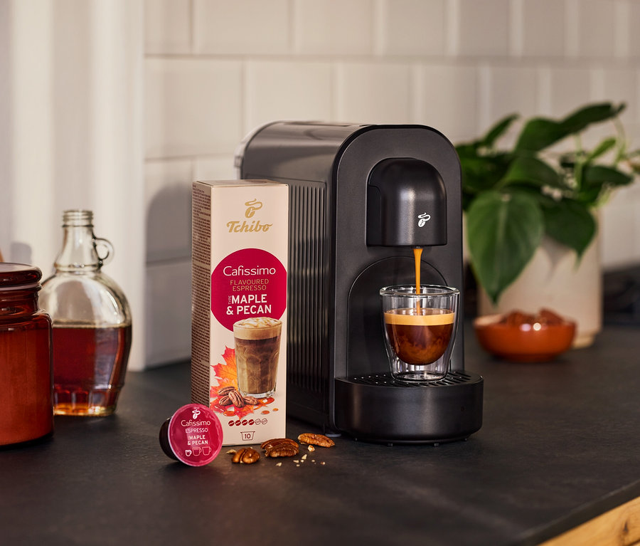 Czarny ekspres do kawy zaparza espresso do szklanki. Obok opakowanie kapsułek o smaku klonowo-orzechowym, luźna kapsułka, orzechy, butelka i roślina.