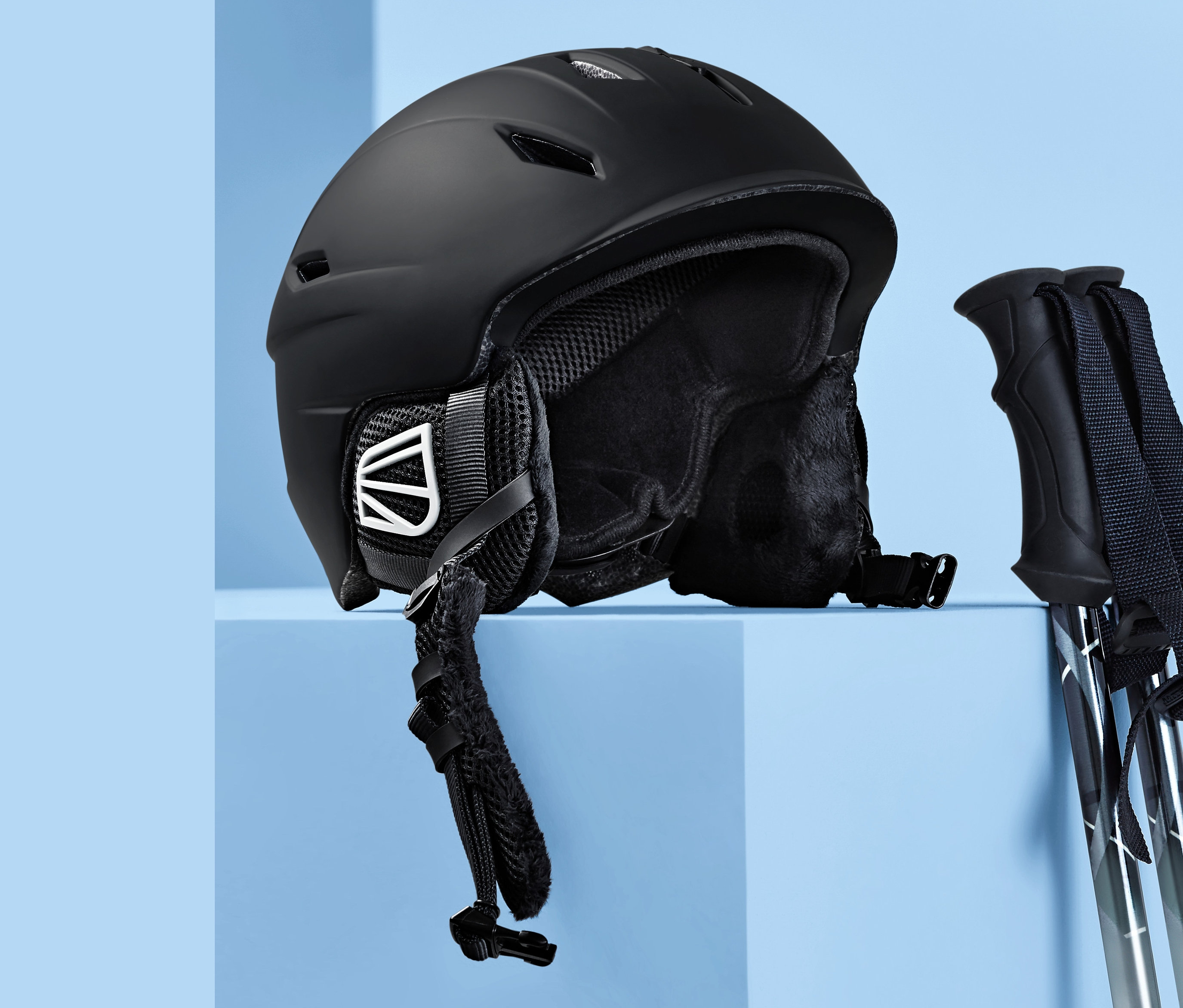 Czarny kask wykonany w technologii in-mould i kijki narciarskie na niebieskim tle.