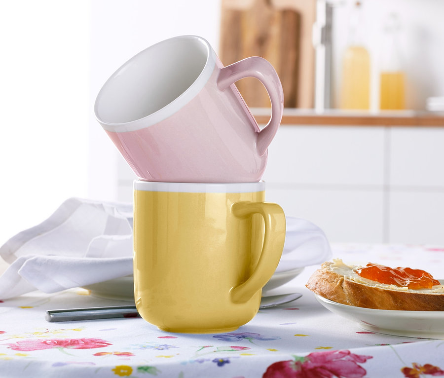 Na stole leżą dwa kubki, jeden różowy i jeden żółty, ułożone jeden na drugim. W tle talerz z chlebem i dżemem.