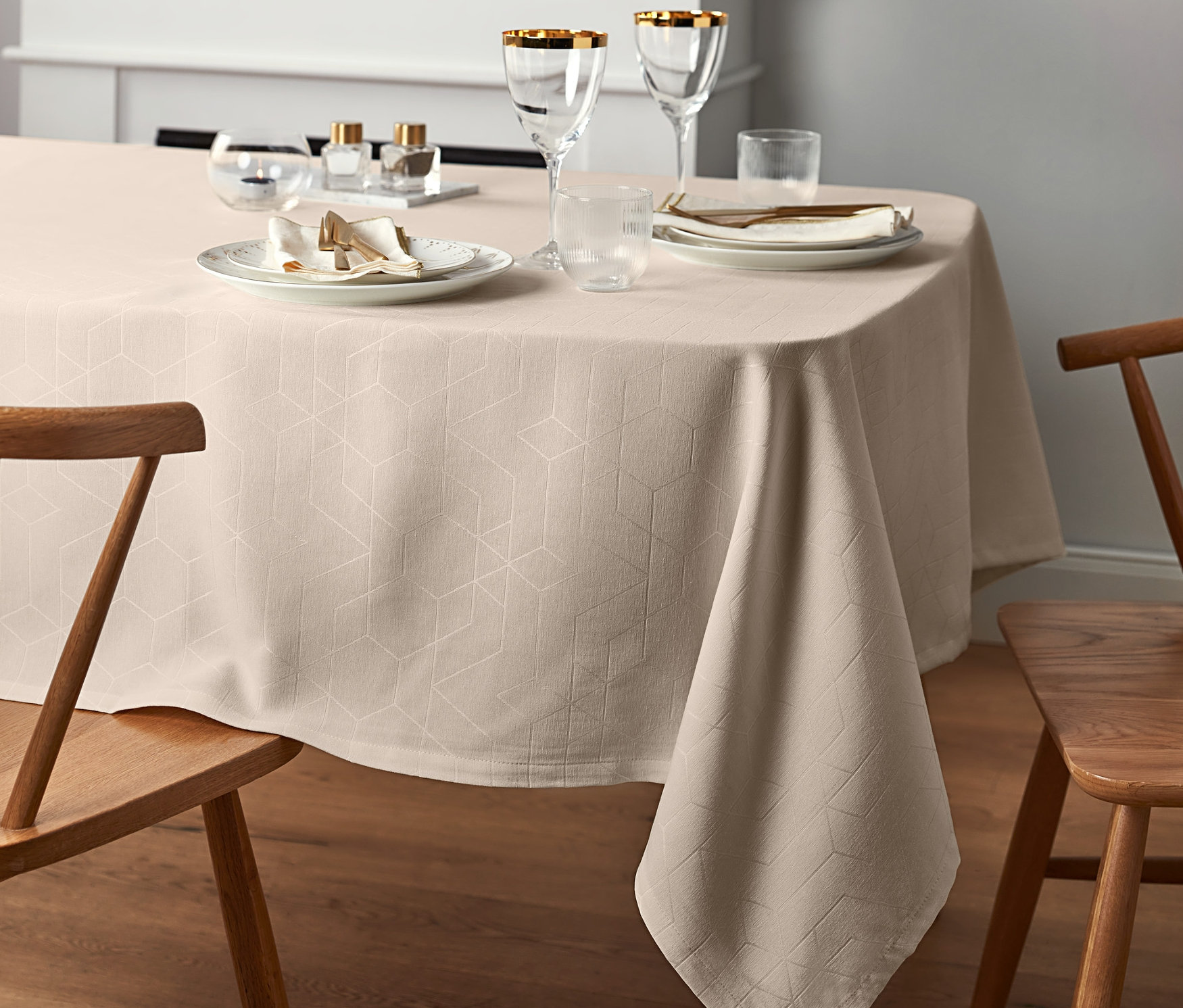Nakryty stół z kremowym obrusem żakardowym o standardowej wielkości, talerzami, szklankami i drewnianymi krzesłami.