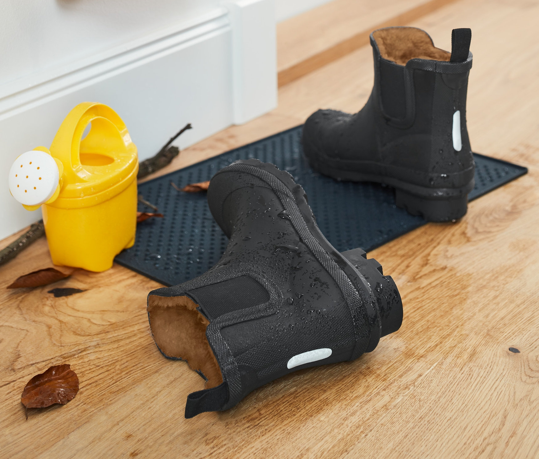 Dwie czarne kalosze z podszewką stoją na niebieskiej wycieraczce przy drzwiach, obok nich żółta konewka i kurtka.