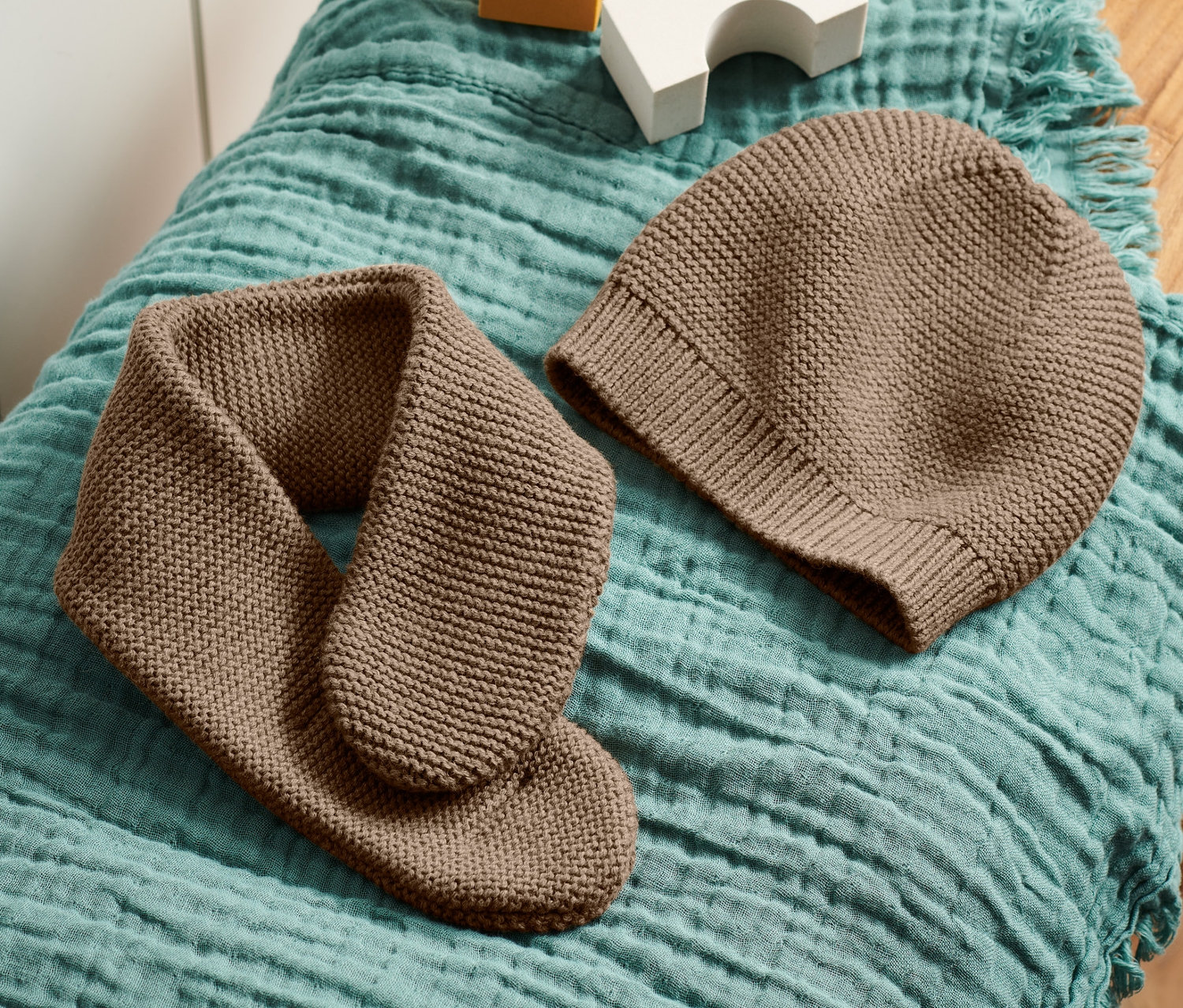 Brązowa czapka i szalik z dzianiny leżą na turkusowym materiale na drewnianym stołku obok białych i żółtych kostek.