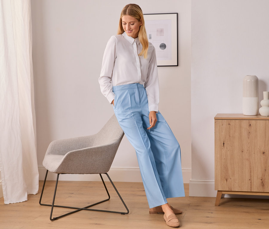 Kobieta w białej koszuli i spodniach z tkaniny w kolorze jasnoniebieskim opiera się o szare krzesło.