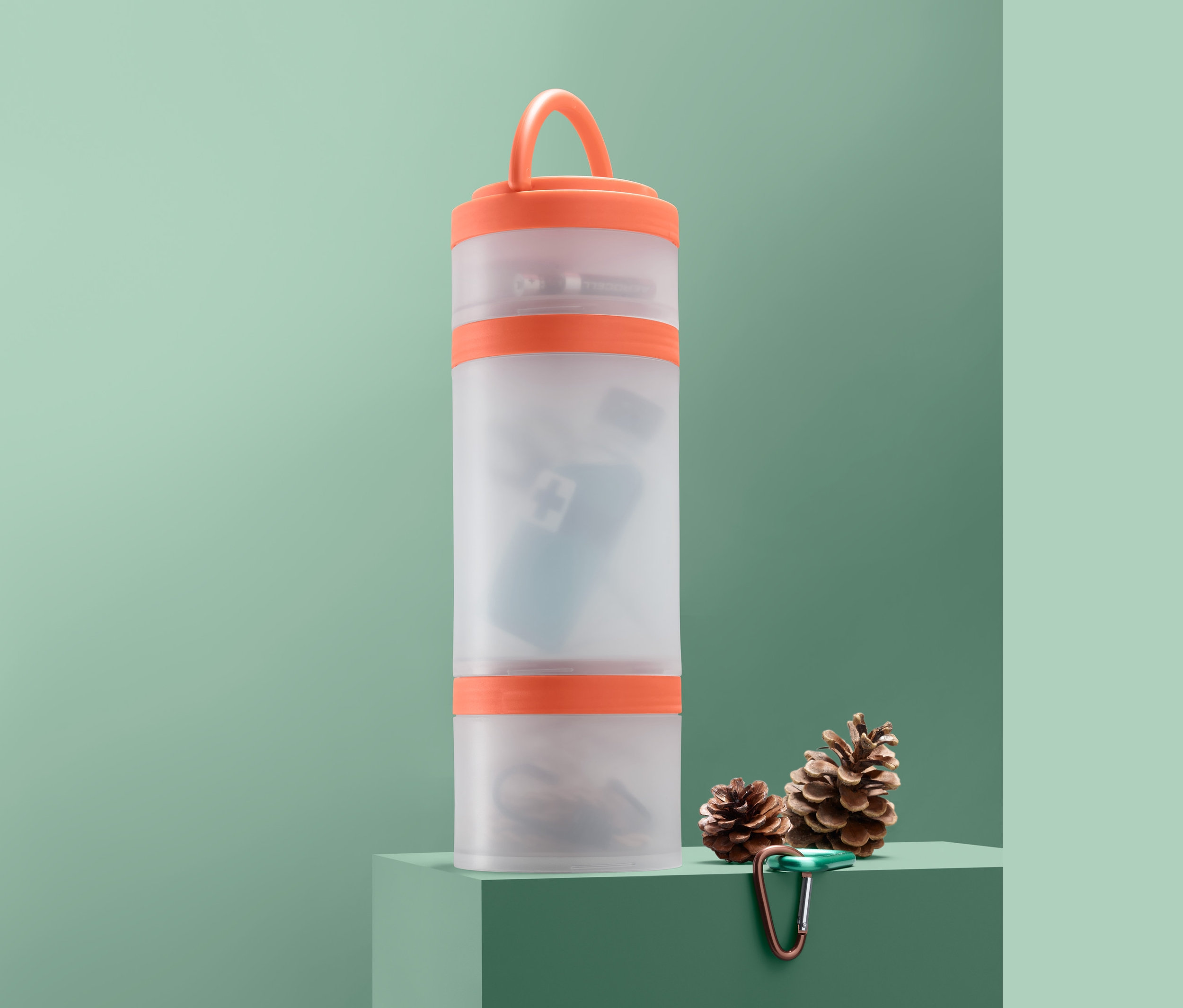 Wielofunkcyjne pojemniki do przechowywania stoją na zielonym cokole obok dwóch szyszek i karabińczyka.