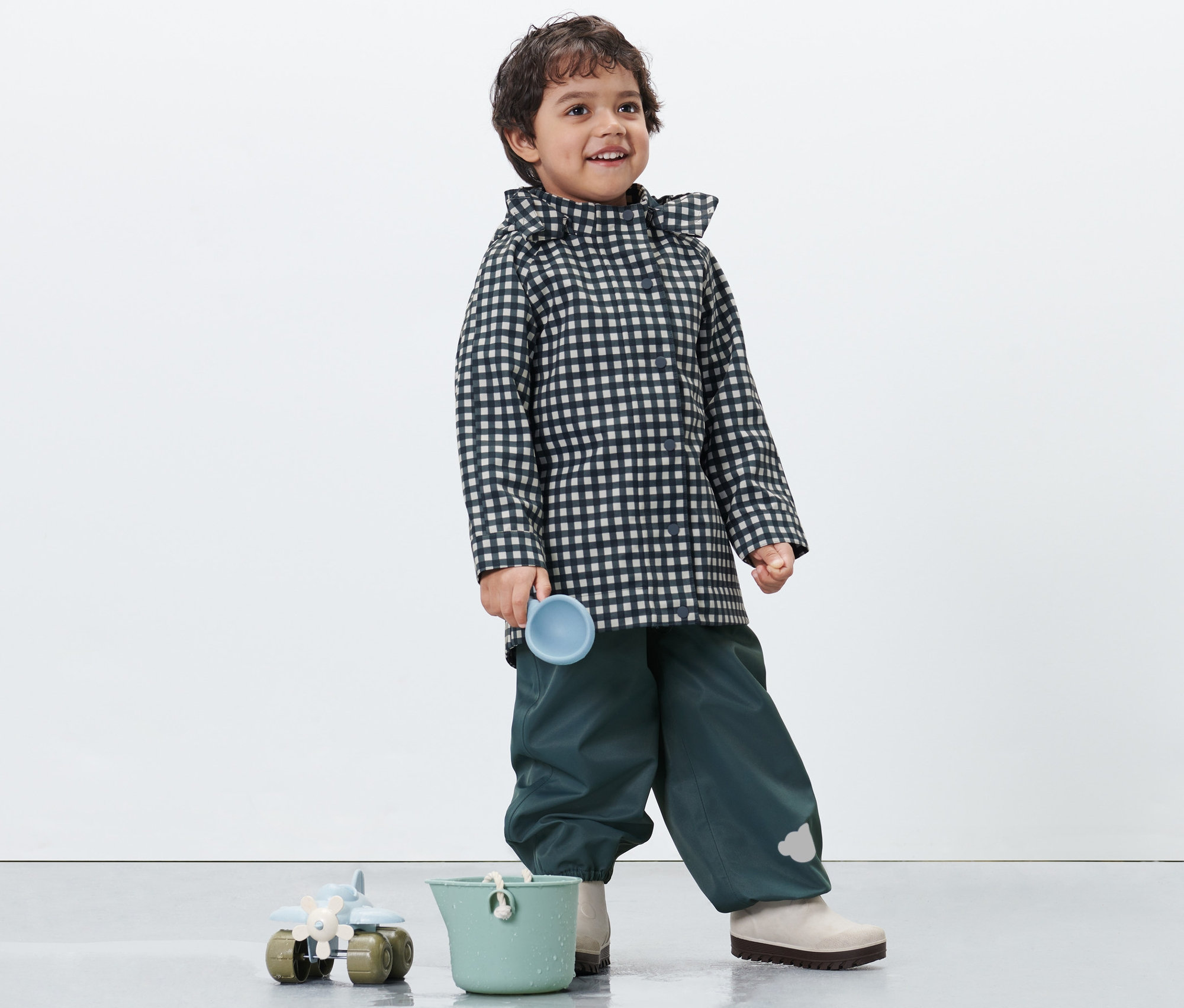 Chłopiec w kurtce przeciwdeszczowej w kratę trzyma niebieski kubek. Na dole stoi zielone wiaderko i zabawka.