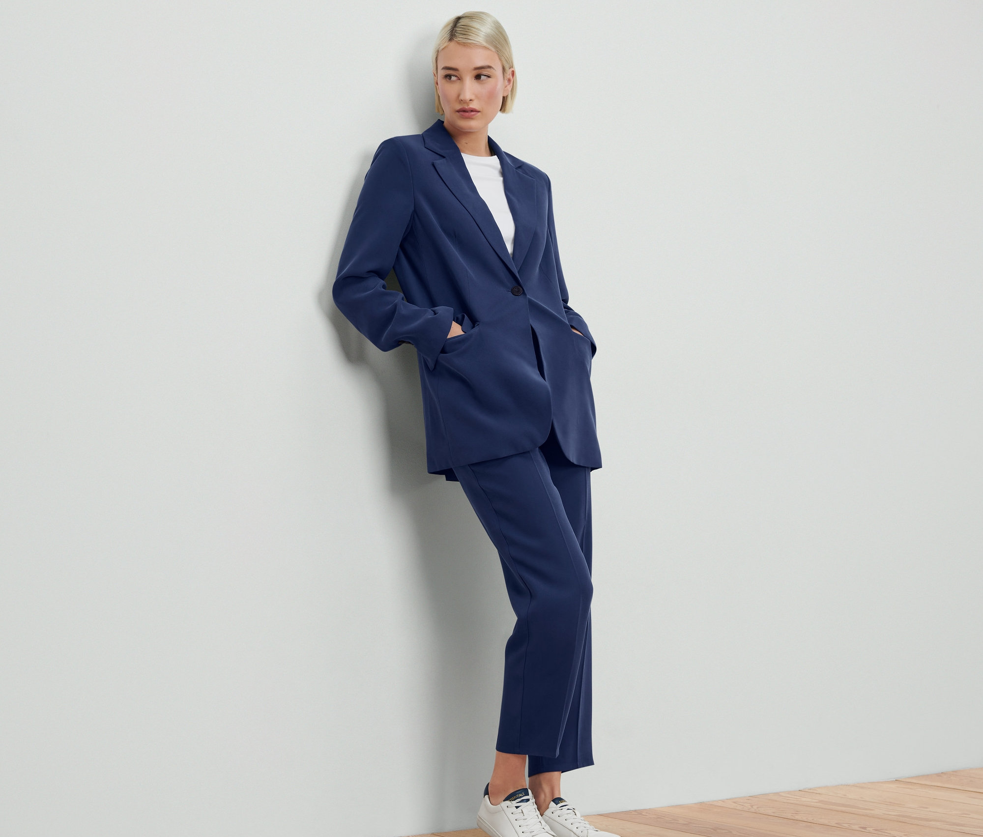 Kobieta opiera się o ścianę, ma na sobie niebieską marynarkę i spodnie, Żakiet z tkaniny.