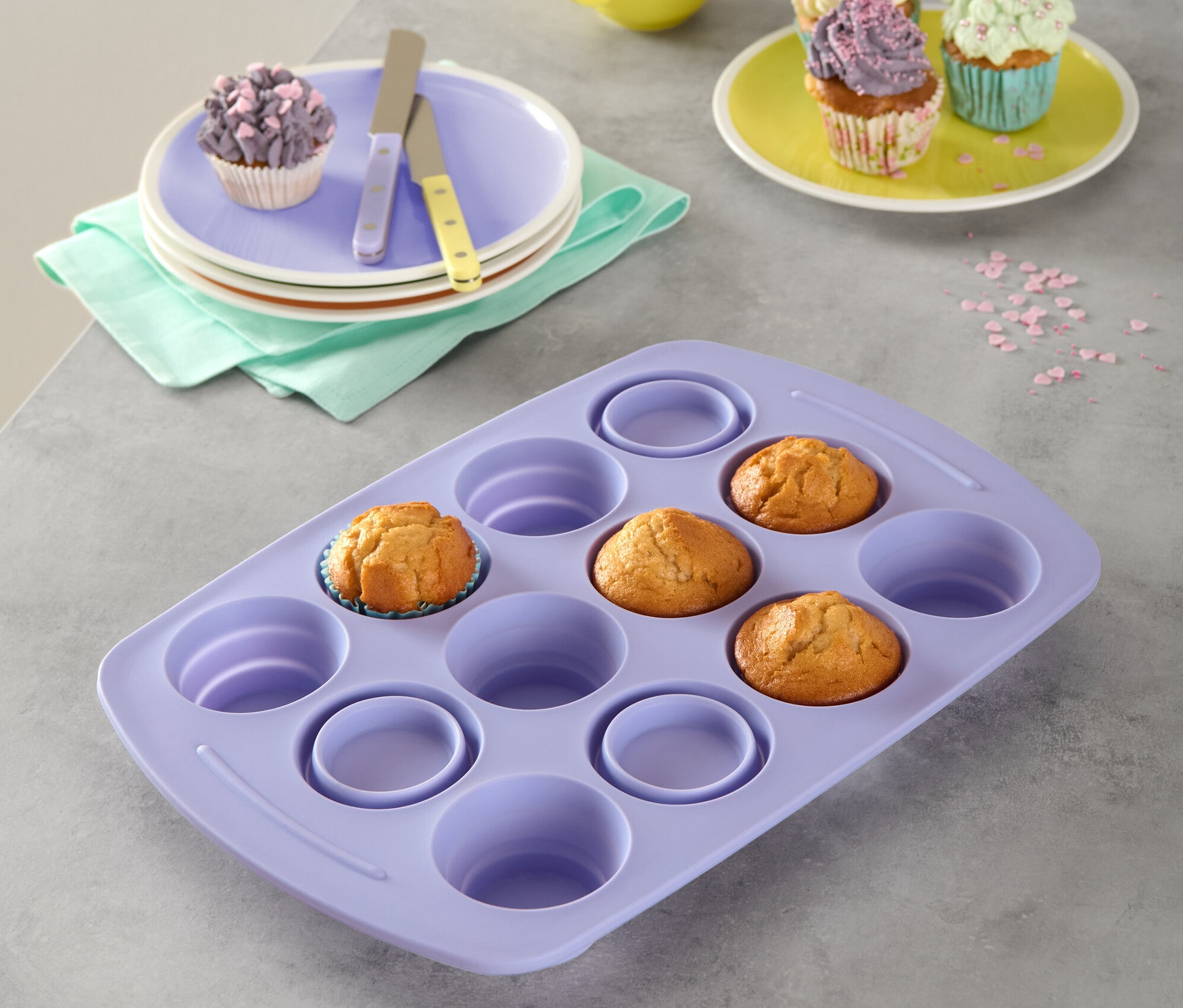 Fioletowa składana silikonowa forma do muffinów z trzema babeczkami. W tle talerze z babeczkami, żółty kubek i posypka.