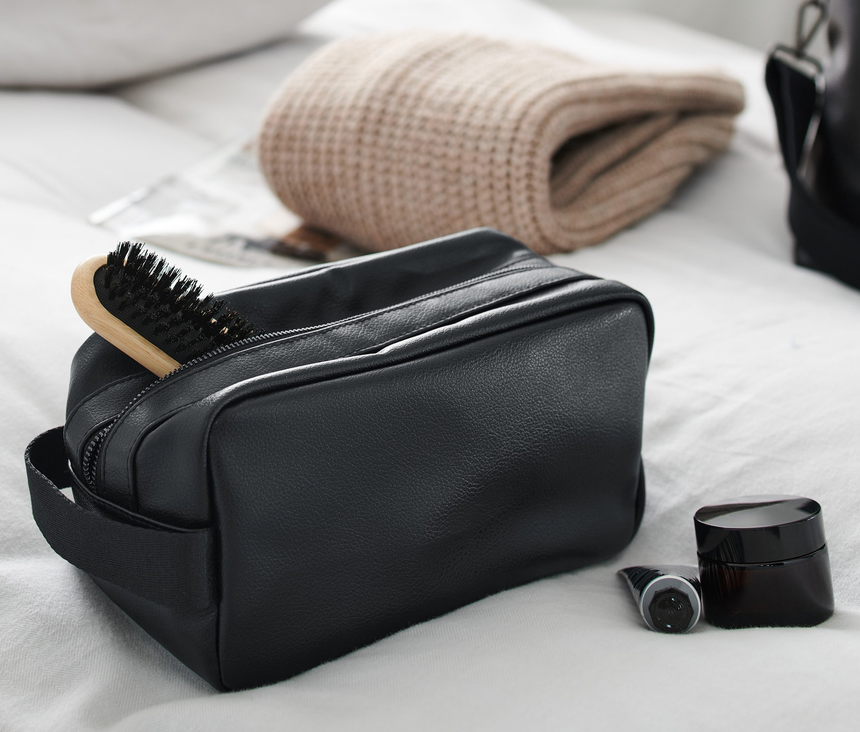 Czarna kosmetyczka z szczotką do włosów, kremem i butelką na białym prześcieradle.