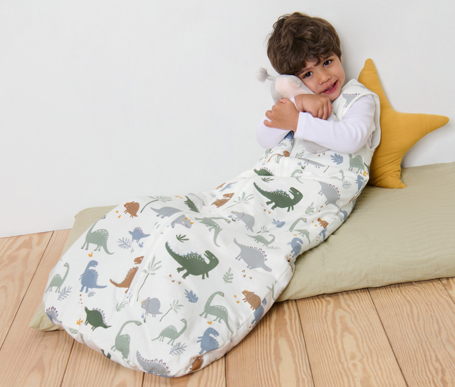 Chłopiec leży w śpiworze 3 TOG, 110 cm we wzór dinozaurów i przytula pluszową zabawkę.