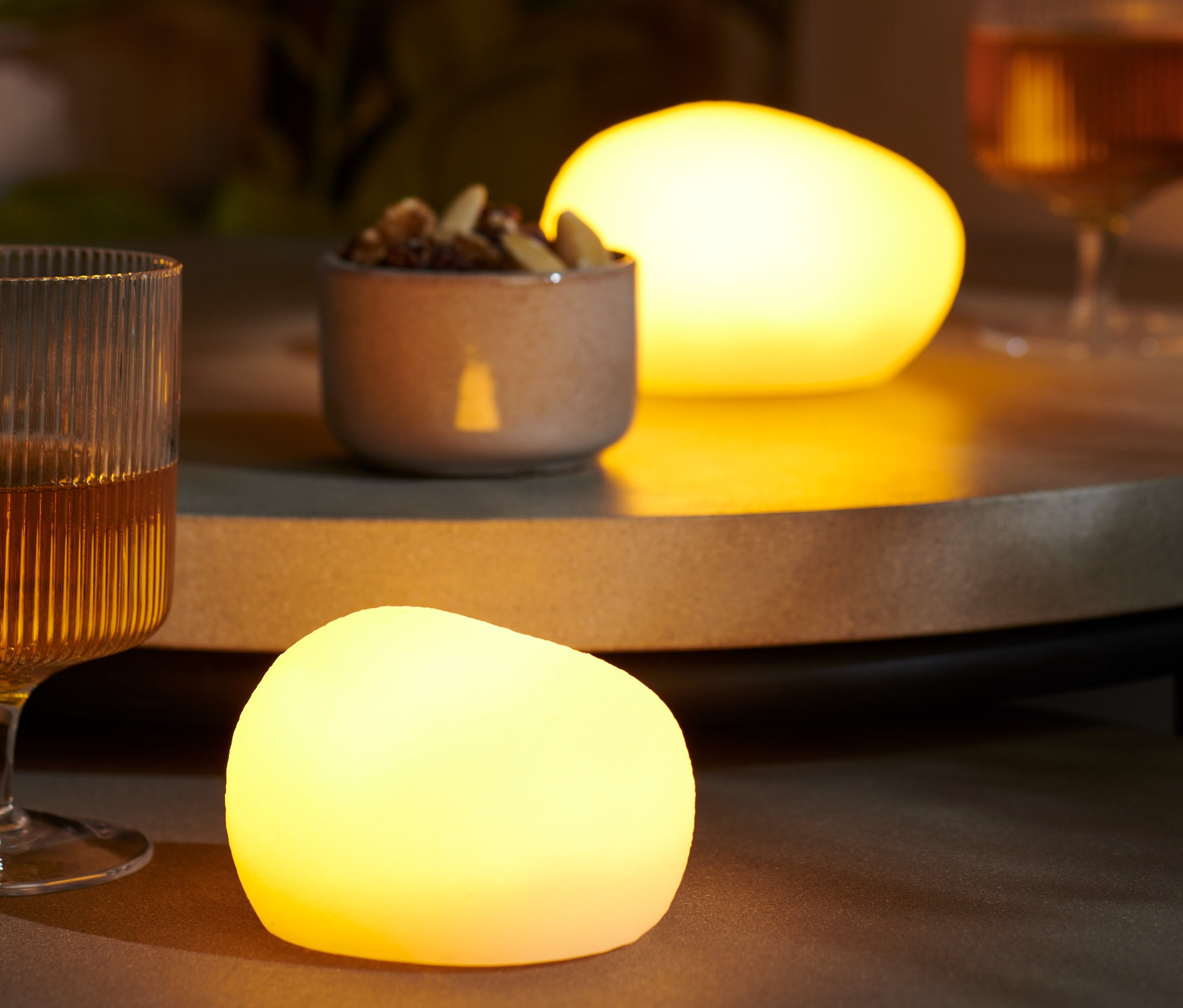 Na stole stoją dwa kamienie dekoracyjne LED, szklanka z napojem i miska orzechów.