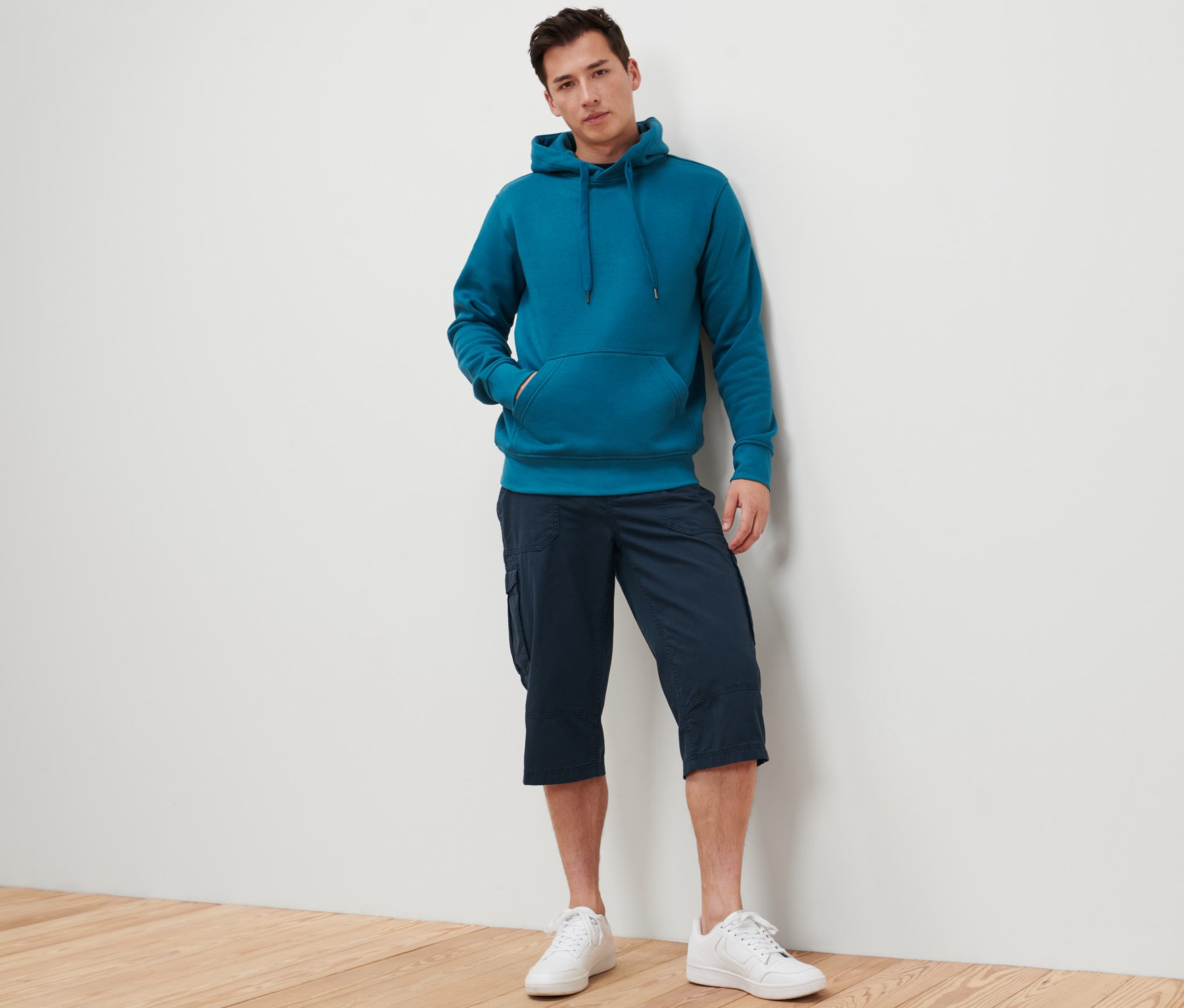 Mężczyzna oparty o ścianę ma na sobie niebiesko-zieloną bluzę z kapturem i granatowe bermudy bojówki o długości 3/4.