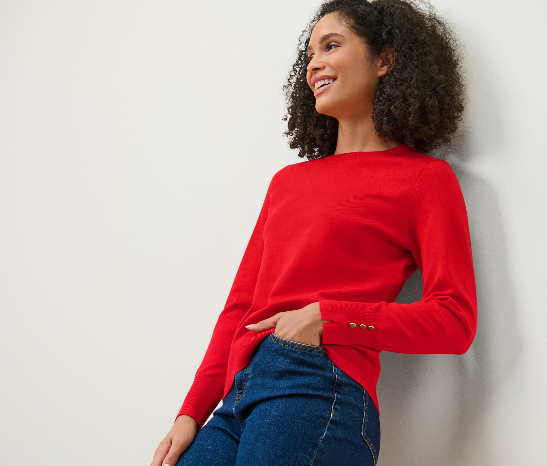 Uśmiechnięta kobieta oparta o ścianę ma na sobie czerwony, cienki sweter i dżinsy.