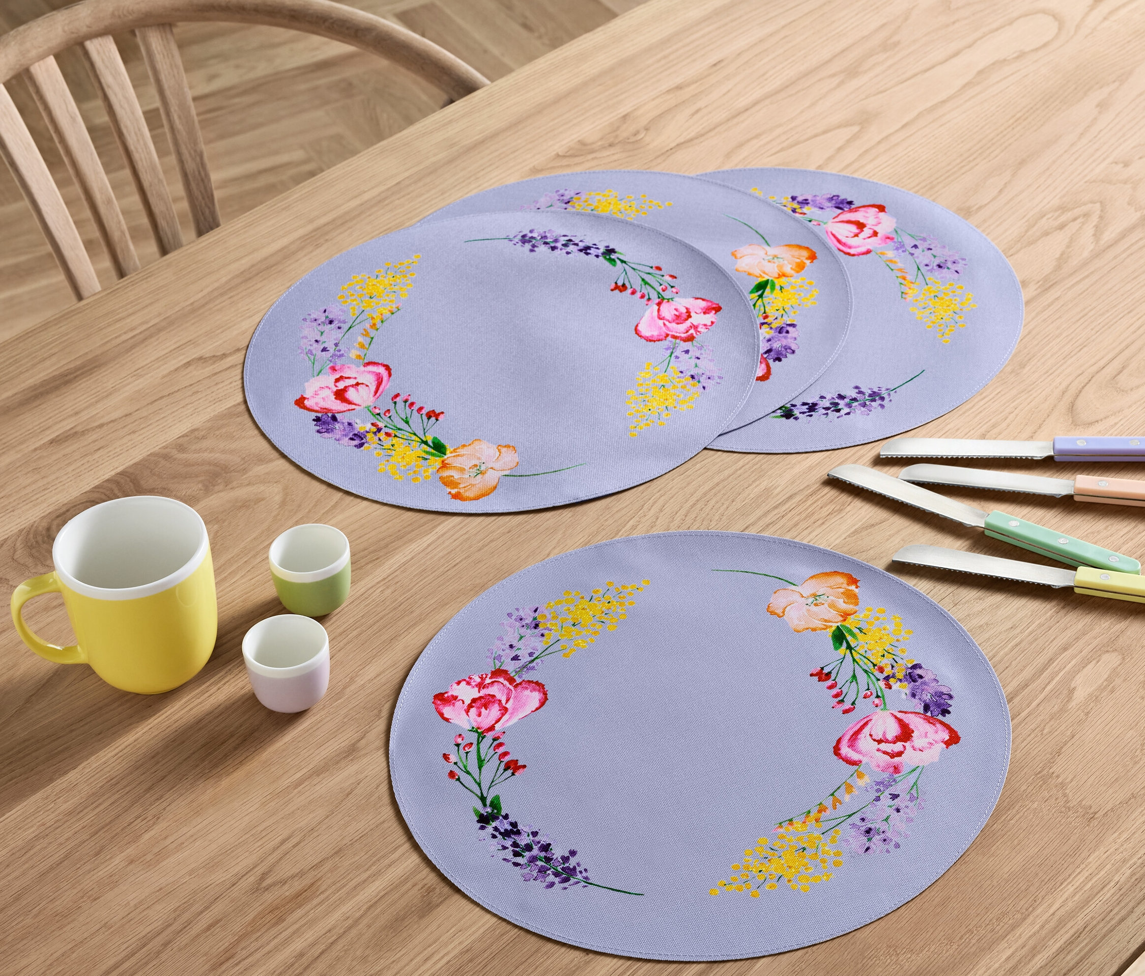 Na drewnianym stole leżą cztery podkładki pod talerze w kwiaty, żółty kubek, dwa małe kubki i cztery noże z kolorowymi rączkami.