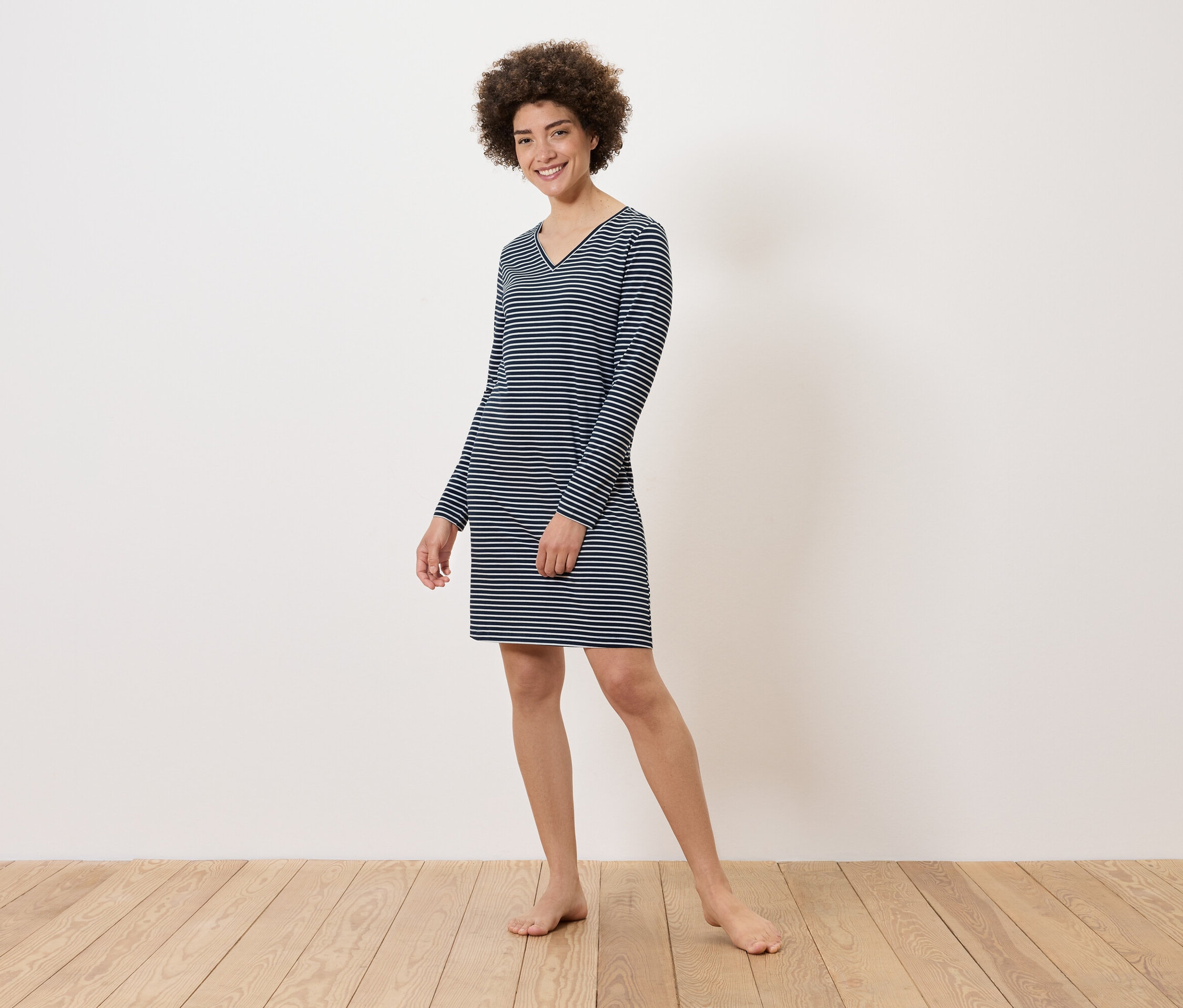 Kobieta stoi boso na drewnianej podłodze, ubrana w sukienkę w paski z długim rękawem.