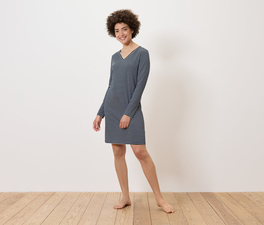 Kobieta stoi boso na drewnianej podłodze, ubrana w sukienkę w paski z długim rękawem.