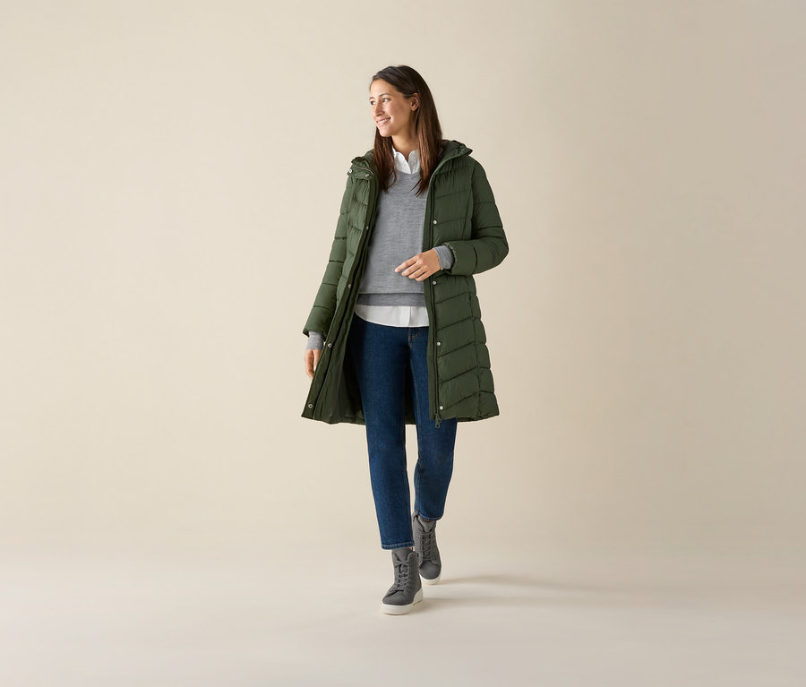 Kobieta ma na sobie szary sweter z cienkiej dzianiny z wełny merino, zieloną kurtkę i dżinsy.