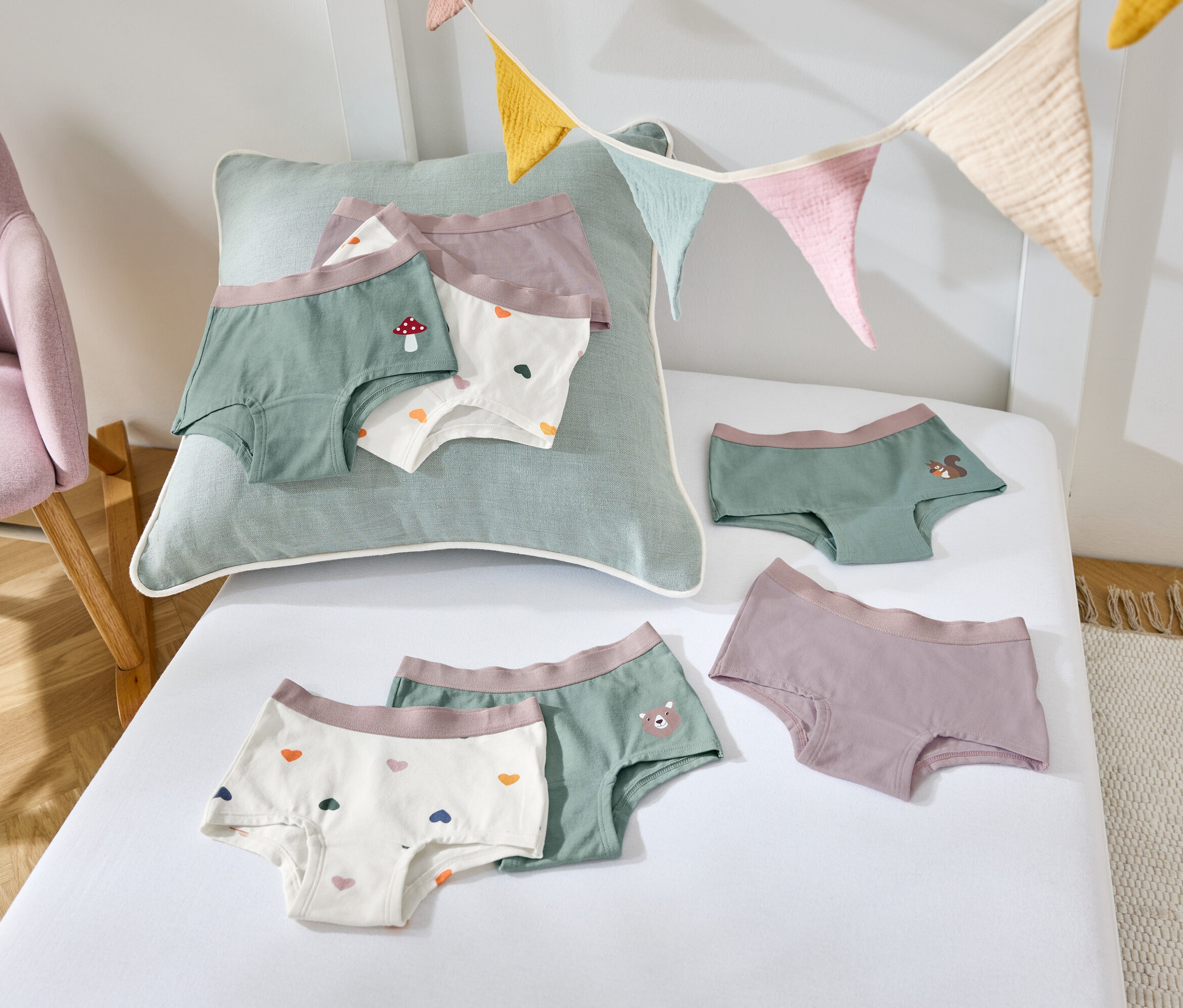 Na białym prześcieradle leżą dziecięce majtki w różnych kolorach i wzorach, obok stoi fioletowy fotel.