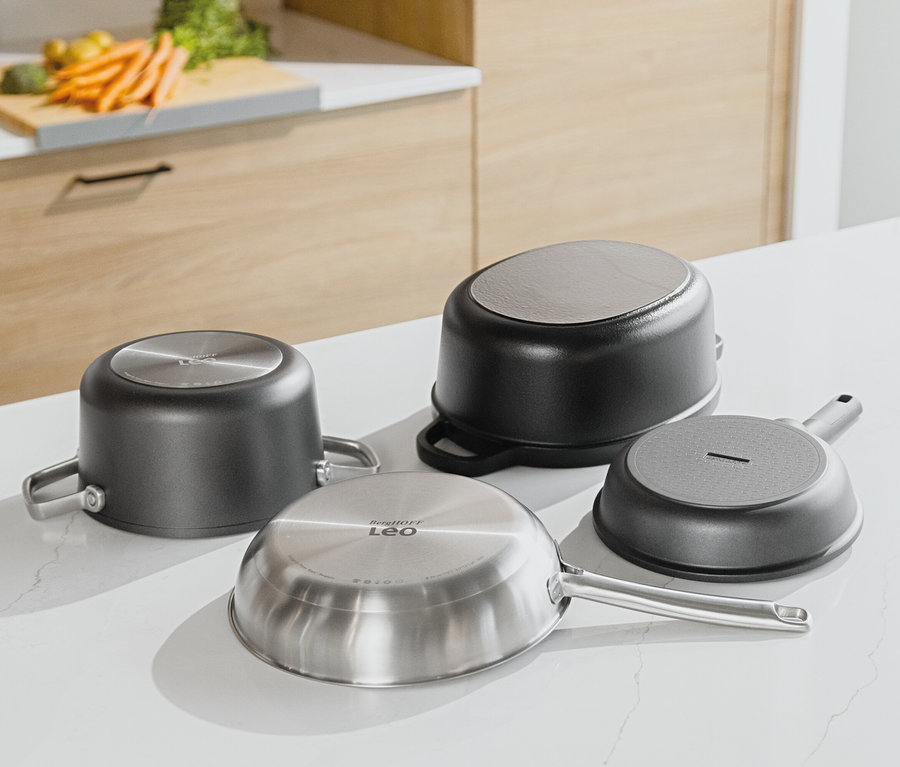 Cztery patelnie leżą dnem do góry na białym blacie kuchennym. W tle widać drewniane szafki kuchenne i warzywa na desce do krojenia.