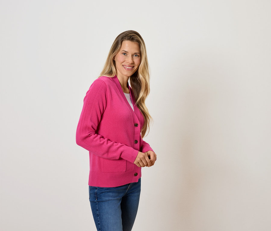 Kobieta z blond włosami pozuje w różowym kardiganie i niebieskich dżinsach.