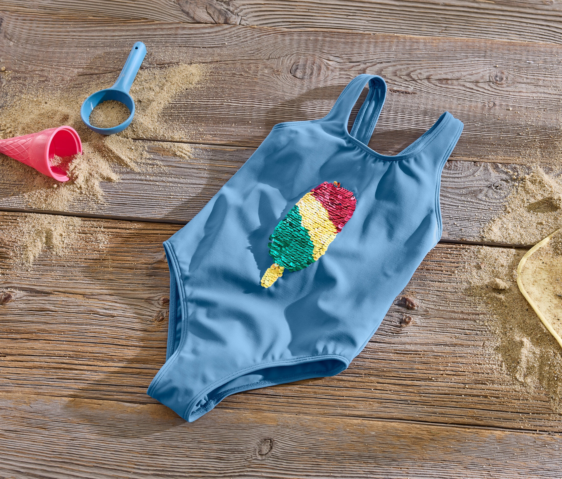 Niebieski kostium kąpielowy z dwustronnymi cekinami leży na drewnianej powierzchni z piaskiem i zabawkami.