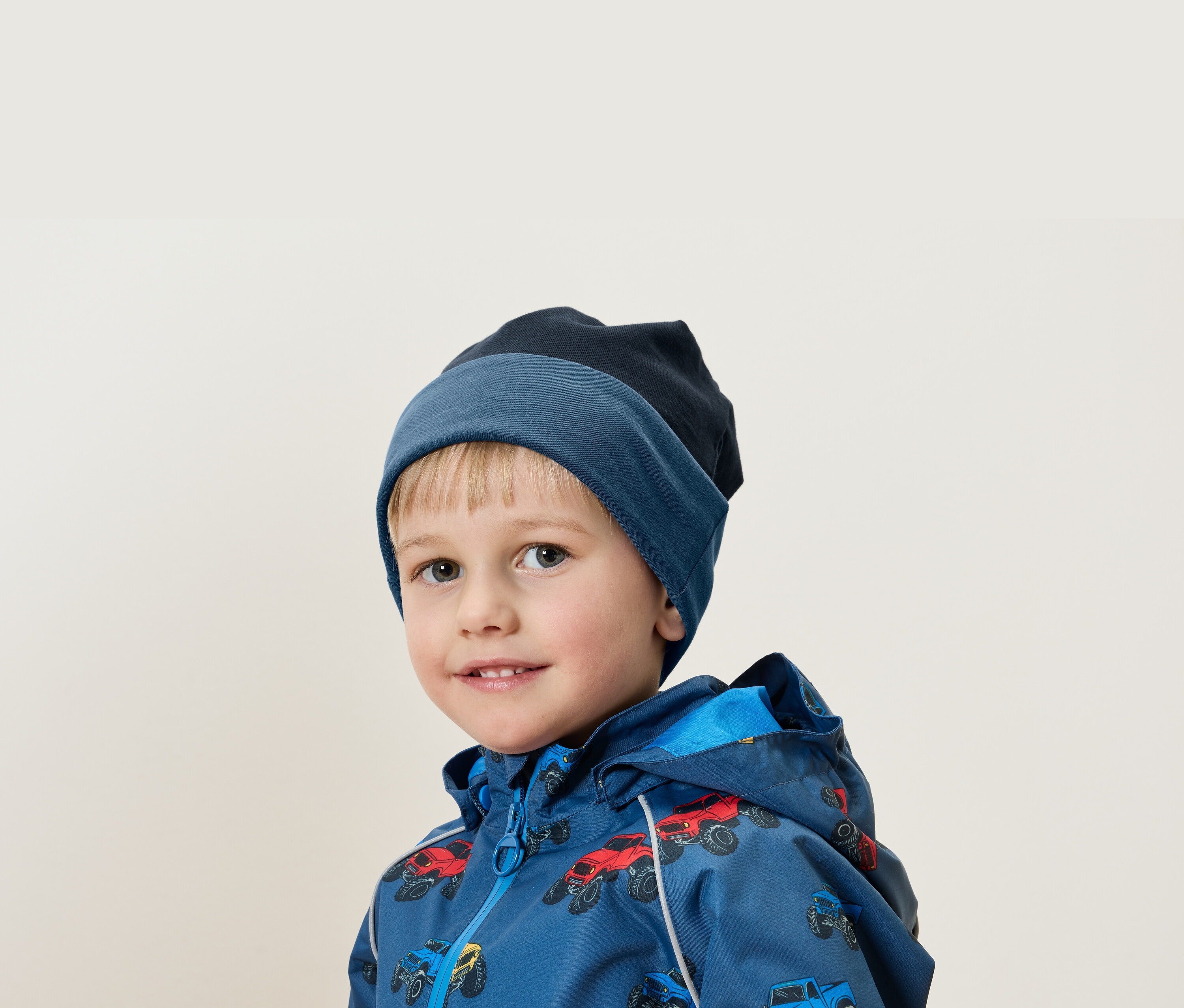 Portret blond chłopca w niebieskiej czapce i kurtce z nadrukiem samochodów.