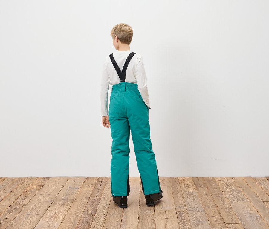 Chłopiec stoi tyłem do kamery, ubrany w turkusowe spodnie narciarskie z czarnymi szelkami.