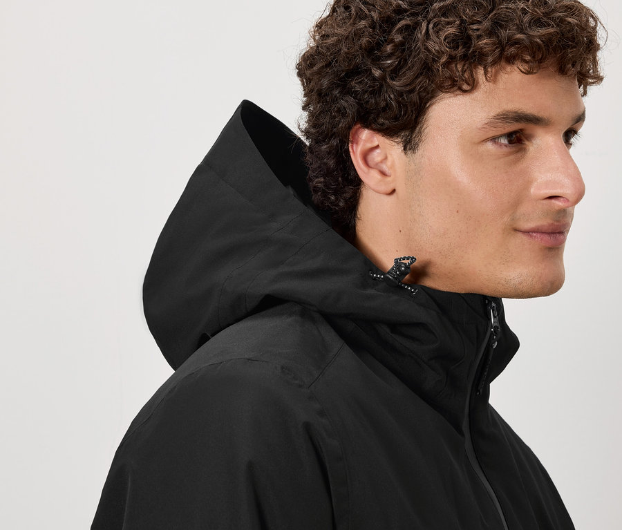 Mężczyzna o brązowych kręconych włosach ma na sobie czarną kurtkę z kapturem.