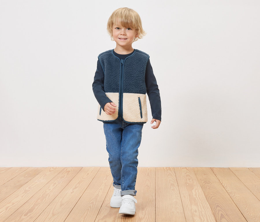 Chłopiec w granatowej kamizelce polarowej z kremowymi panelami na dole zapinanej na zamek stoi na drewnianej podłodze.