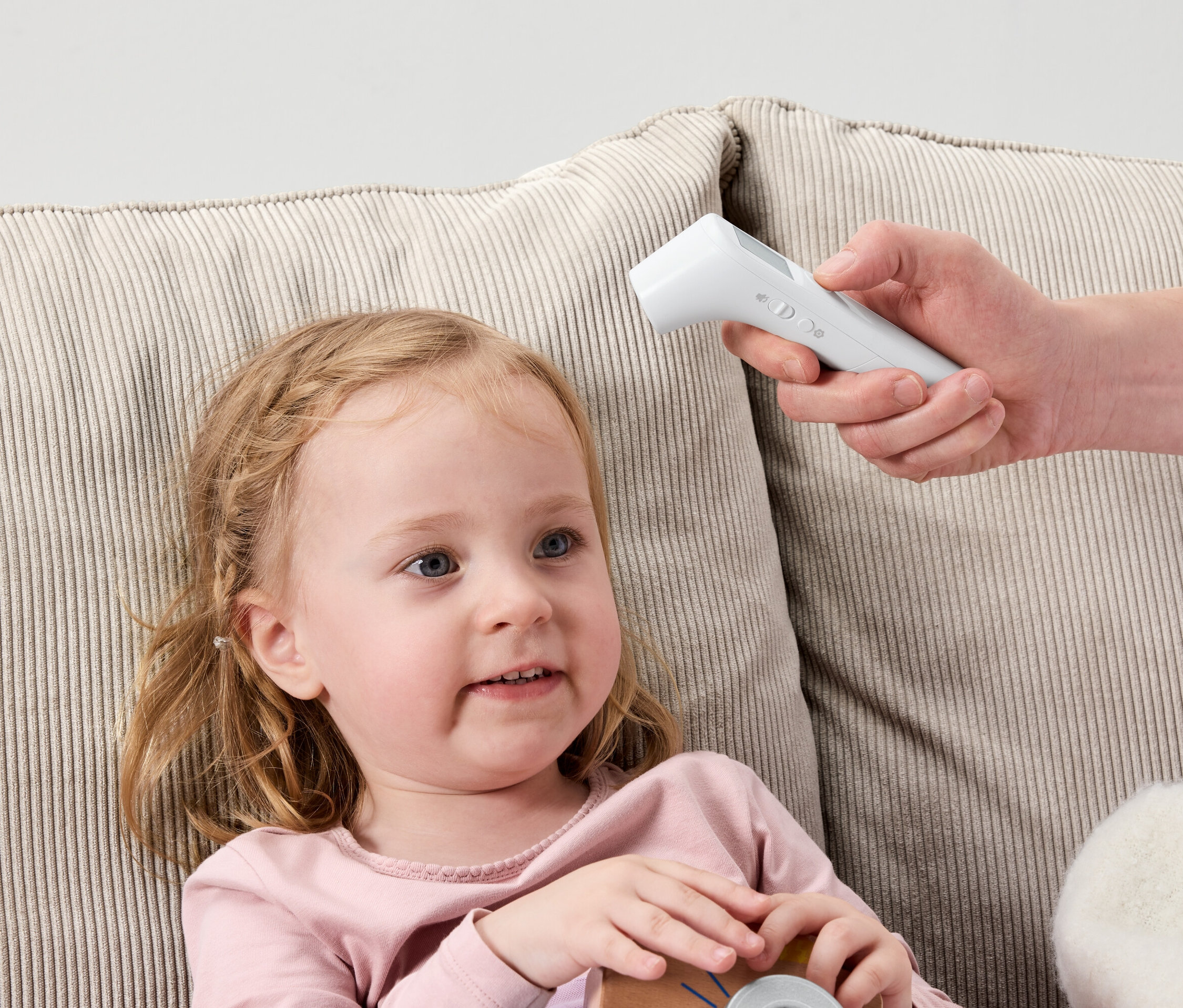 Mała dziewczynka leży na sofie, a ręka trzyma biały termometr cyfrowy przy jej czole.