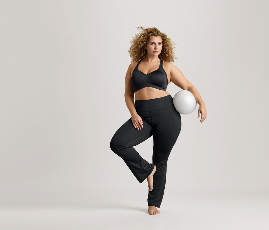 Kobieta o kręconych włosach balansuje na jednej nodze, trzyma białą piłkę, ma na sobie czarny biustonosz sportowy i czarne legginsy.
