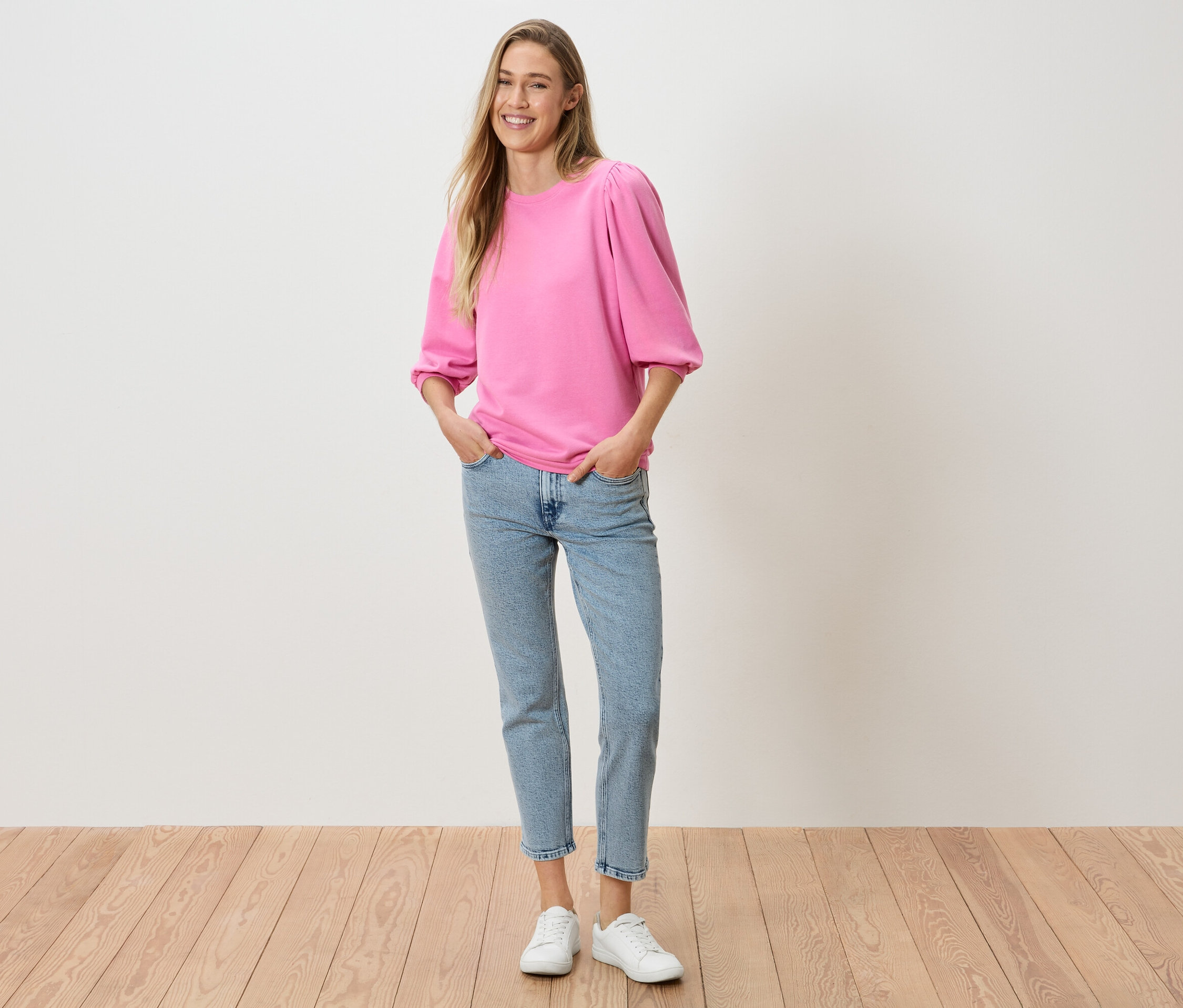 Kobieta z rękami w kieszeniach spodni, ubrana w różową bluzkę z bufiastymi rękawami, dżinsy i białe tenisówki.