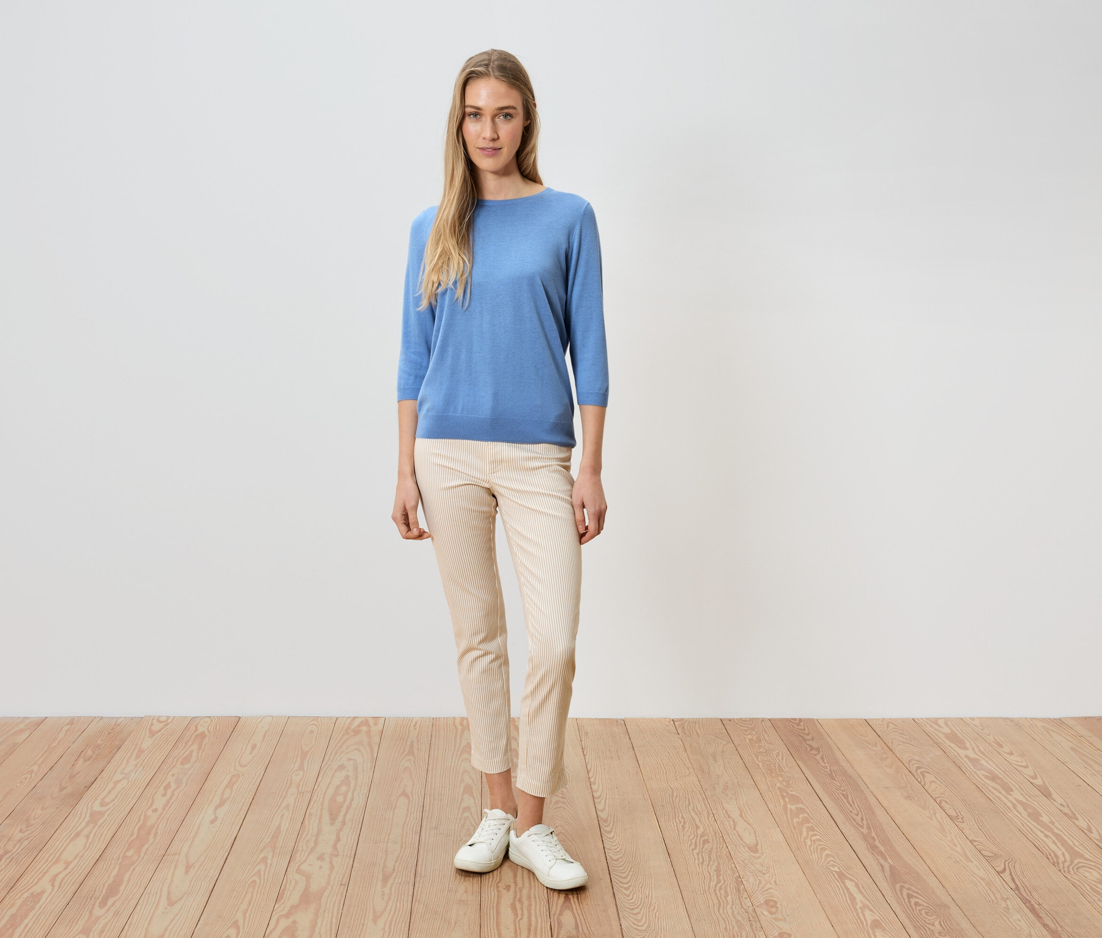 Kobieta stoi na drewnianej podłodze na tle białej ściany, ubrana w niebieski sweter, jasne spodnie i białe buty.