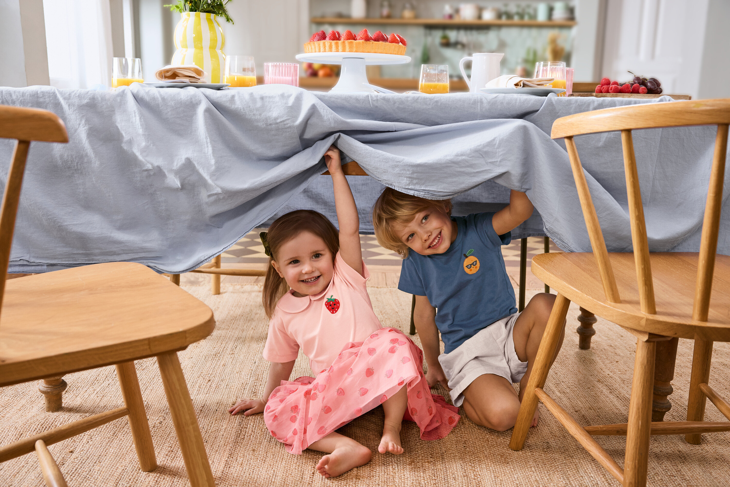 Dwoje dzieci siedzi pod stołem z jasnoniebieskim obrusem. Dziewczynka ma na sobie Dziecięcą spódniczkę tiulową i 2 Dziecięce T-shirty. Chłopiec ma na sobie Dziecięce szorty.