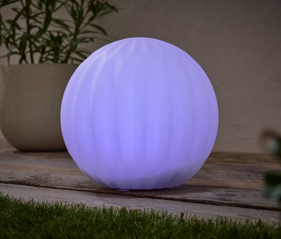 Okrągła, fioletowa lampa stoi na drewnianym stole obok rośliny doniczkowej.