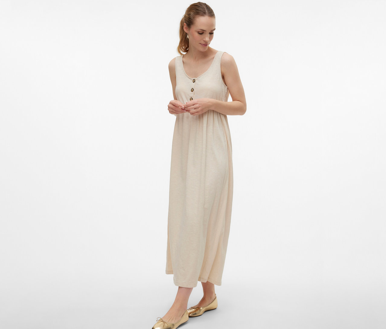 Kobieta w sukience ciążowej MAMALICIOUS „MLEVI LIA” z guzikami i złotych balerinach na białym tle.