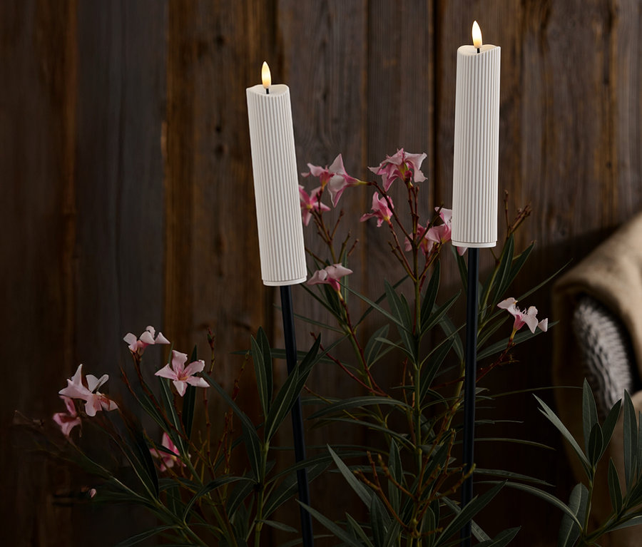 Dwie białe, żebrowane świece na czarnych stojakach pośród różowych kwiatów i zielonych liści na tle ciemnego drewnianego tła.