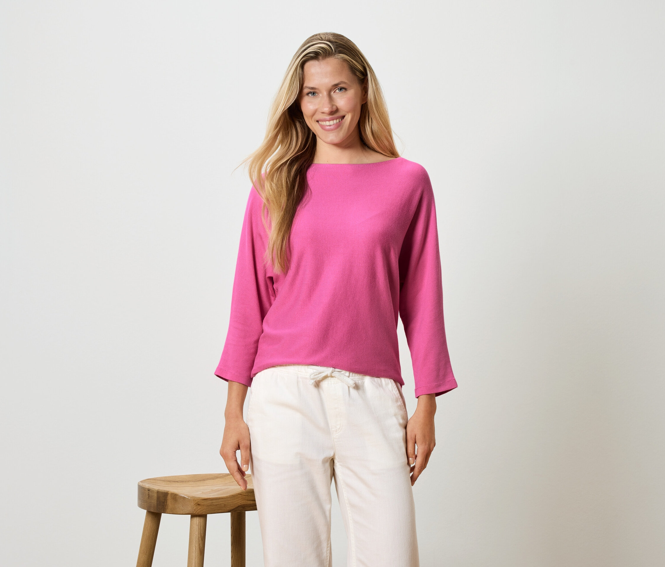 Kobieta z długimi blond włosami ma na sobie różową bluzkę z długim rękawem i jasne spodnie, stoi obok stołka.