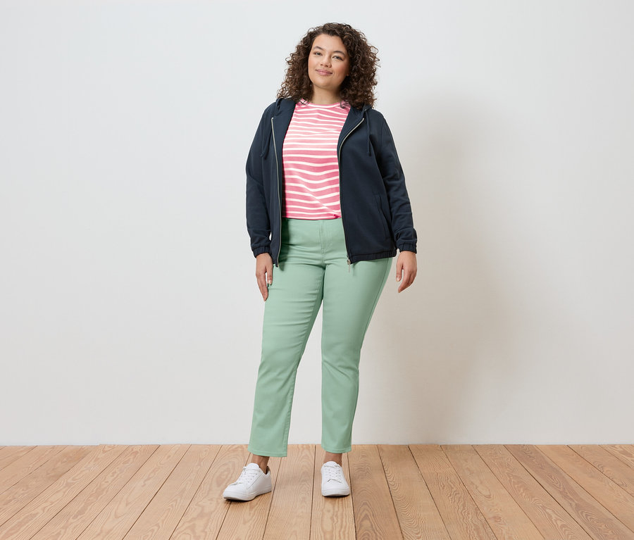 Kobieta stoi w jasnym otoczeniu, ubrana w granatową kurtkę, bluzkę w paski, jasnozielone spodnie i białe tenisówki.
