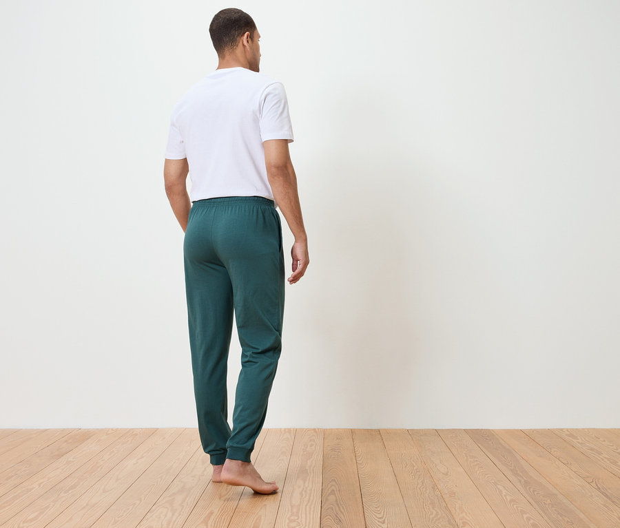 Mężczyzna od tyłu w białej koszulce i zielonych spodniach dresowych, stojący na drewnianej podłodze przed białą ścianą.