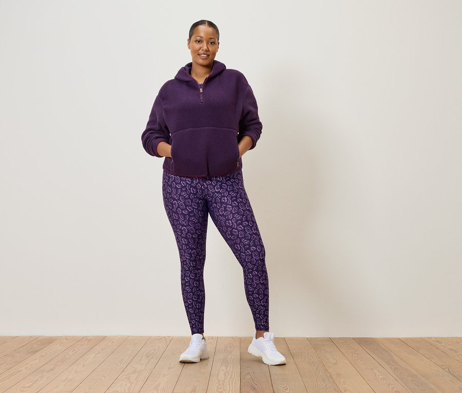 Kobieta w fioletowej bluzie polarowej zapinanej na pół zamek i fioletowych legginsach w cętki lamparta, ubrana w białe tenisówki.