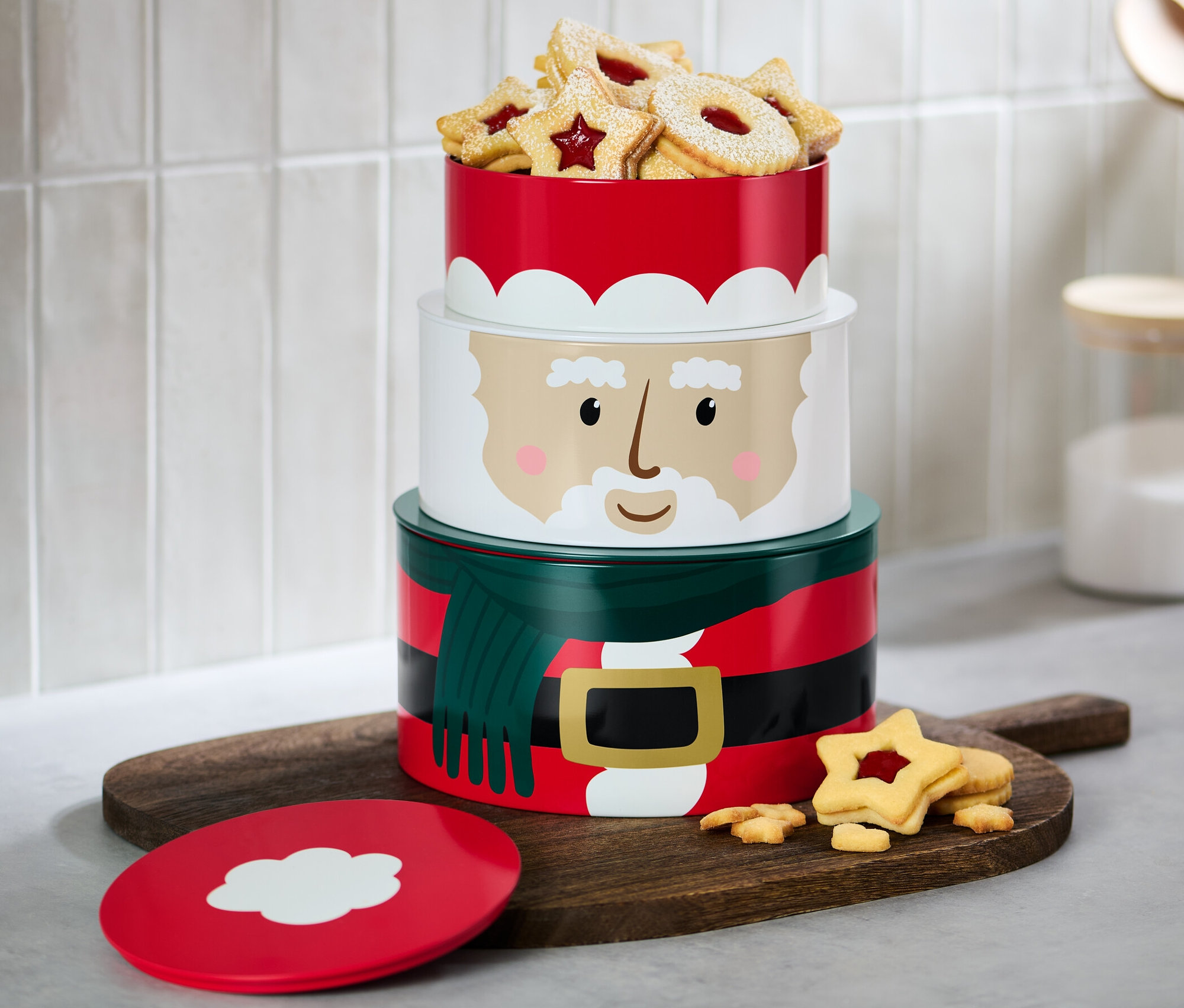 Trzypoziomowe puszki na ciastka z motywem świątecznym wypełnione ciastkami stoją na drewnianej desce.