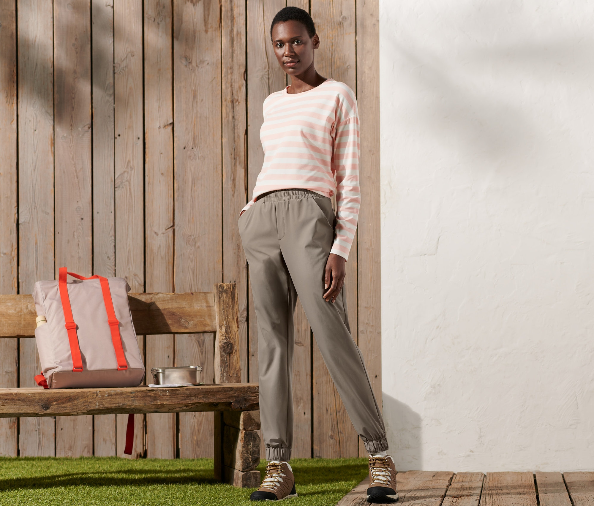 Kobieta stoi i pozuje na drewnianej podłodze. Ma na sobie spodnie funkcyjne i koszulę w paski.