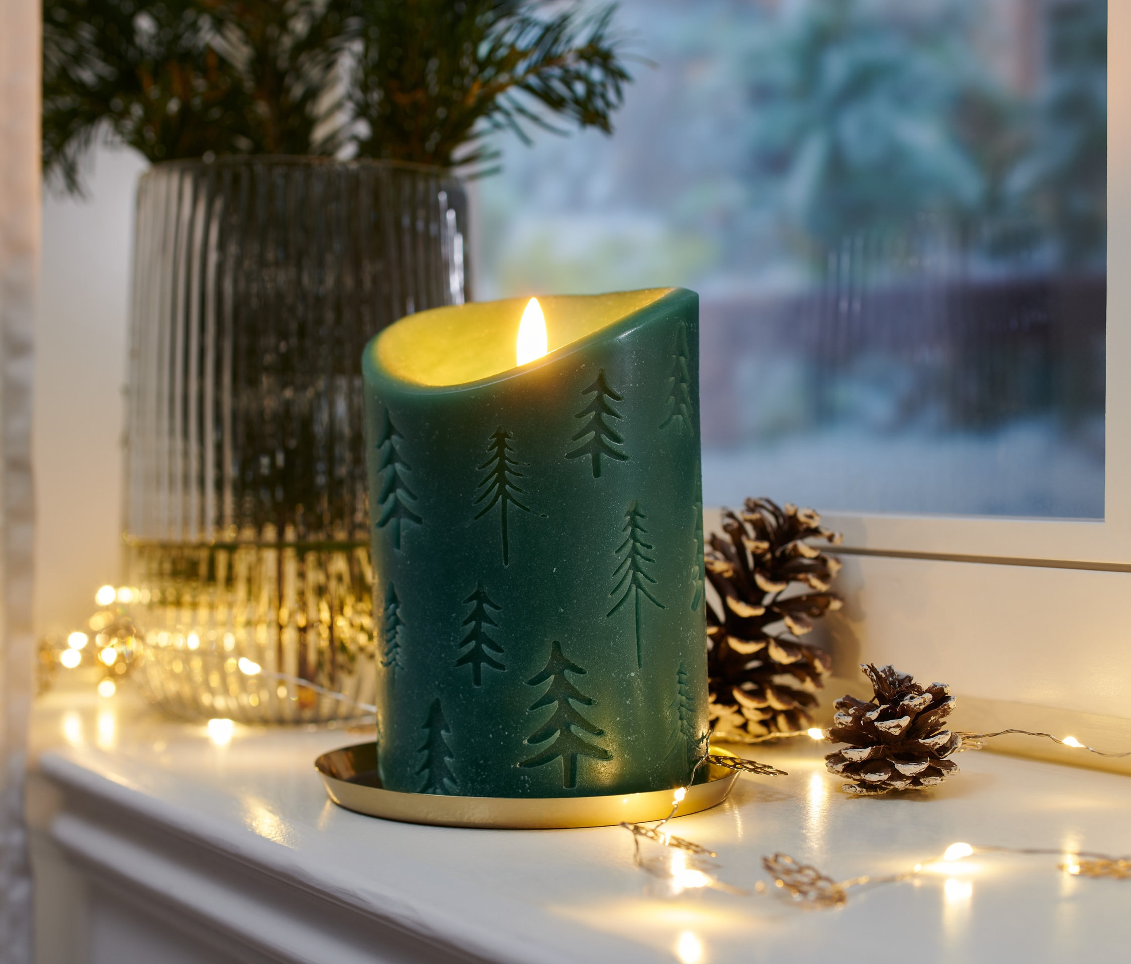 Zielona świeca LED z prawdziwego wosku ze wzorem choinki na złotej tacy obok szyszek i światełek.