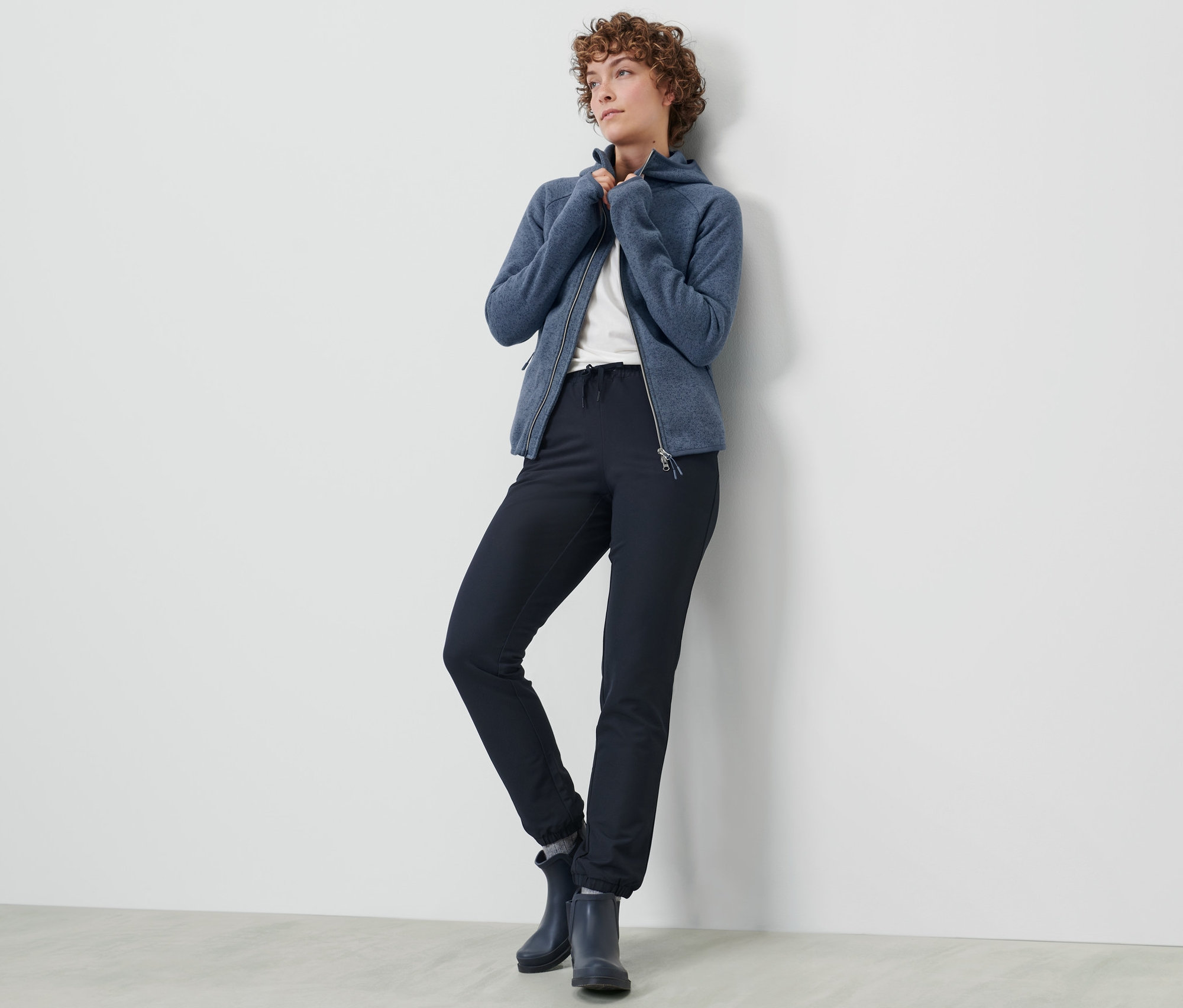 Kobieta pozuje w spodniach dresowych z softshellu i niebieskiej bluzie na zamek błyskawiczny przy ścianie.