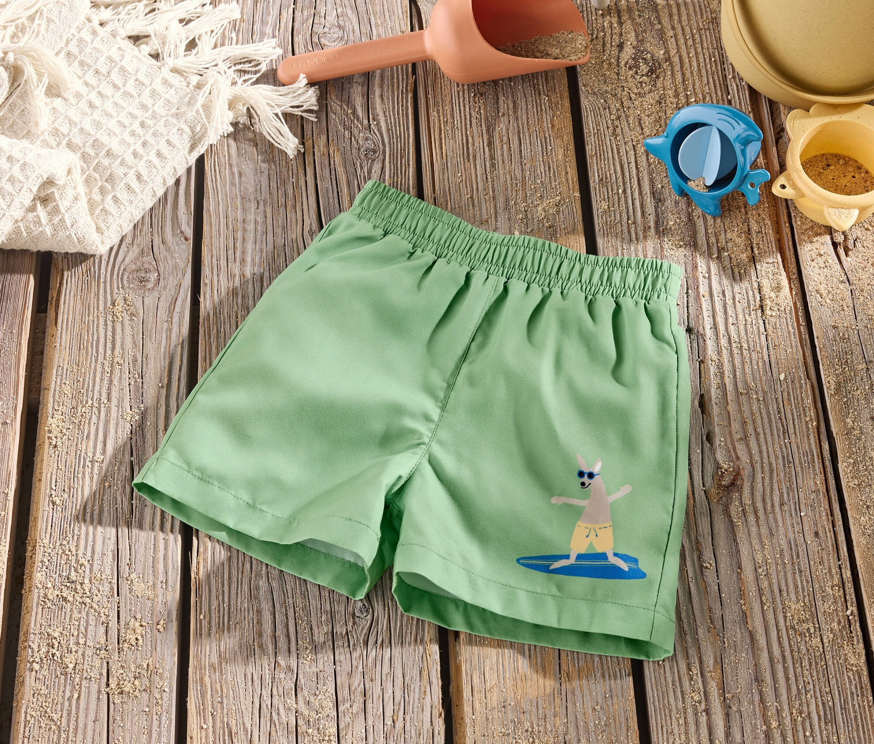 Zielone szorty kąpielowe z kangurem na desce surfingowej leżą na drewnianym stole z zabawkami do piasku.