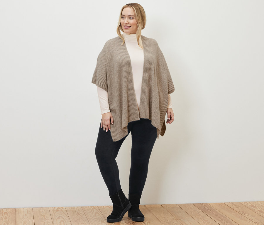 Kobieta ma na sobie ponczo z dzianiny, sztruksowe legginsy i kremowo-białą bluzkę z kołnierzem golfowym.