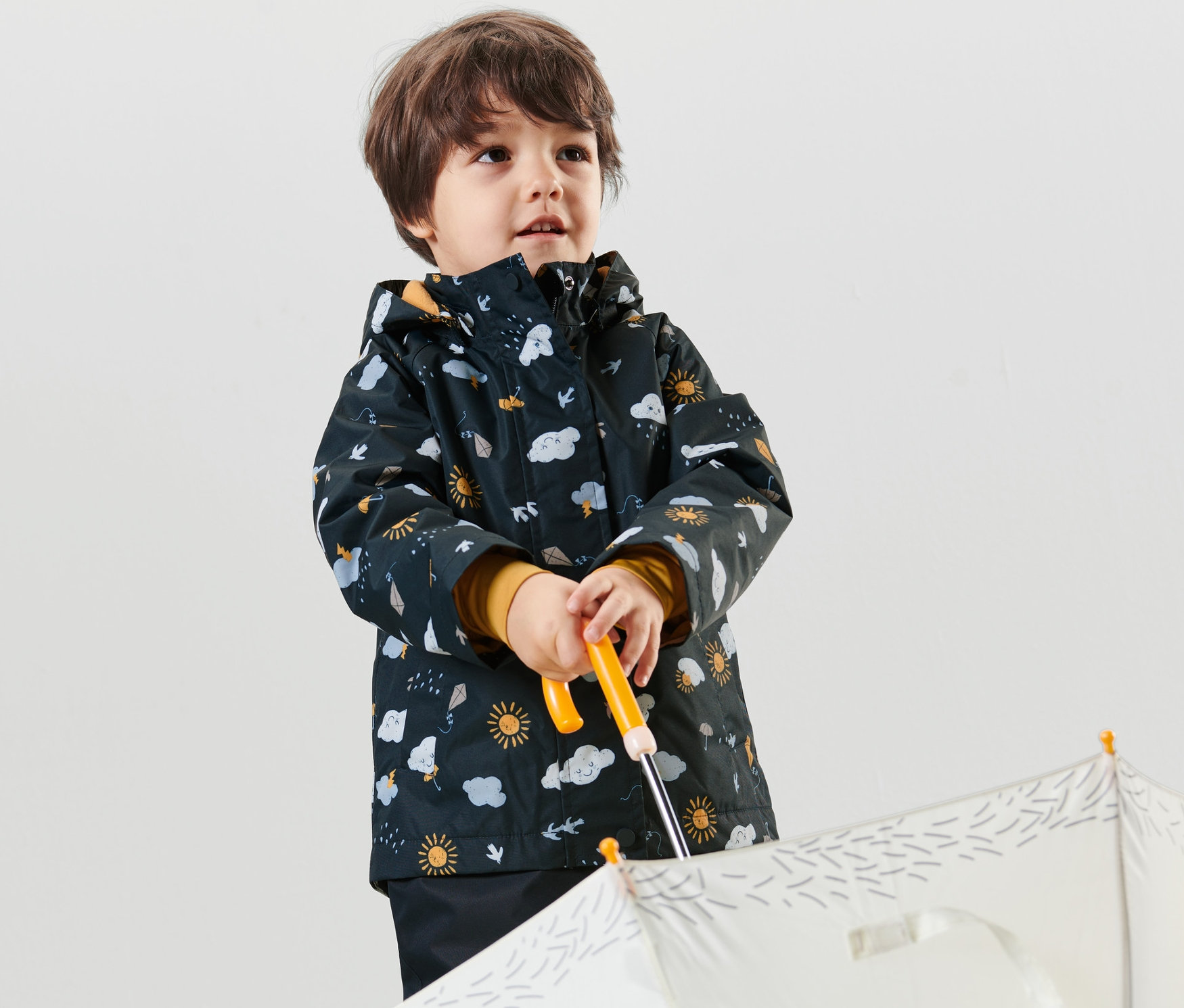 Chłopiec trzyma biały parasol i ma na sobie granatową, dziecięcą termiczną kurtkę przeciwdeszczową z nadrukiem na całej powierzchni.